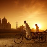 India, Uttar Pradesh, AgraMan on bike with girl on trailer and man pushing from behind in front of Taj Mahal at sunset