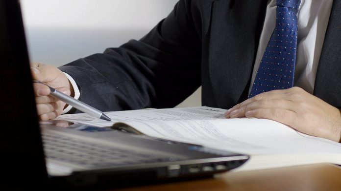 businessman-at-work-with-laptop-is-checking-a-printed-list-on-paper-sheets_person_CEO-job