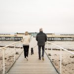 Full length rear view of senior couple walking with Schnauzer on boardwalk at beach against clear sky